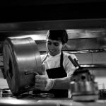 Chef despeja o conteúdo de uma panela em uma cozinha profissional movimentada.