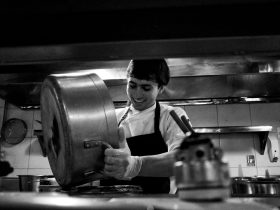 Chef despeja o conteúdo de uma panela em uma cozinha profissional movimentada.