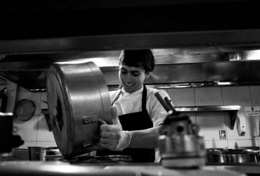 Chef despeja o conteúdo de uma panela em uma cozinha profissional movimentada.