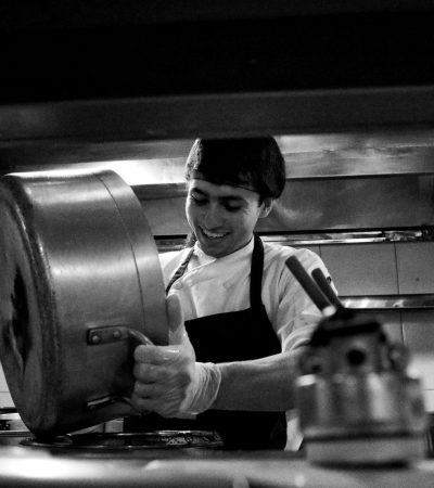 Chef despeja o conteúdo de uma panela em uma cozinha profissional movimentada.