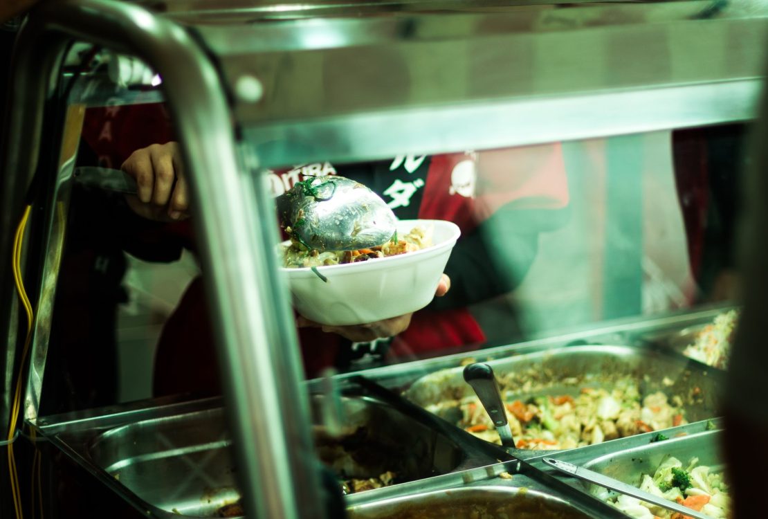 Pessoa servindo pratos variados em um balcão de comida brasileira.