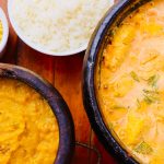Moqueca brasileira colorida servida em panela de barro, acompanhada de guarnições sobre uma mesa de madeira.