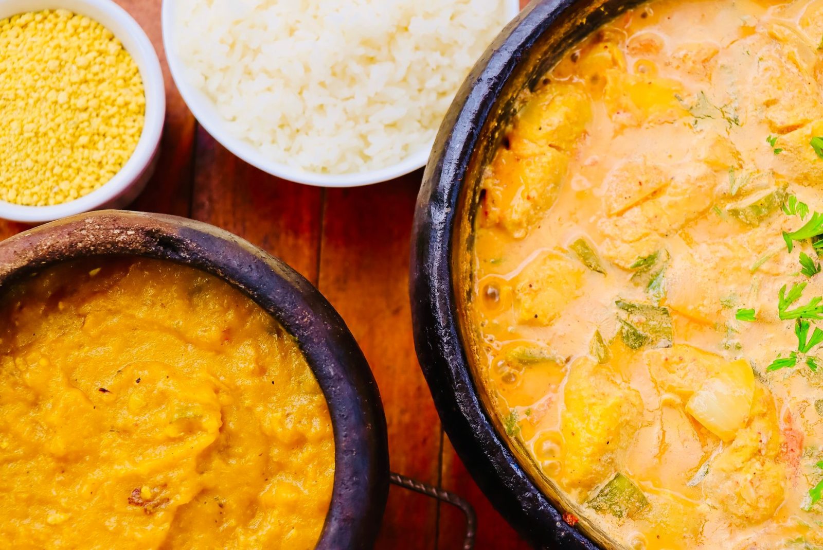 Moqueca brasileira colorida servida em panela de barro, acompanhada de guarnições sobre uma mesa de madeira.