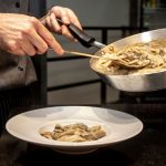 Chef finaliza a montagem de um prato de massa cremosa com cogumelos em uma cozinha.