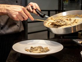 Chef finaliza a montagem de um prato de massa cremosa com cogumelos em uma cozinha.