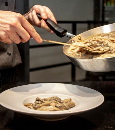 Chef finaliza a montagem de um prato de massa cremosa com cogumelos em uma cozinha.